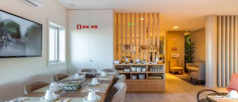 Un espace repas moderne avec une longue table en bois dressée pour un repas, un coin cuisine équipé de divers plats et ustensiles, et un coin salon avec des fauteuils confortables.