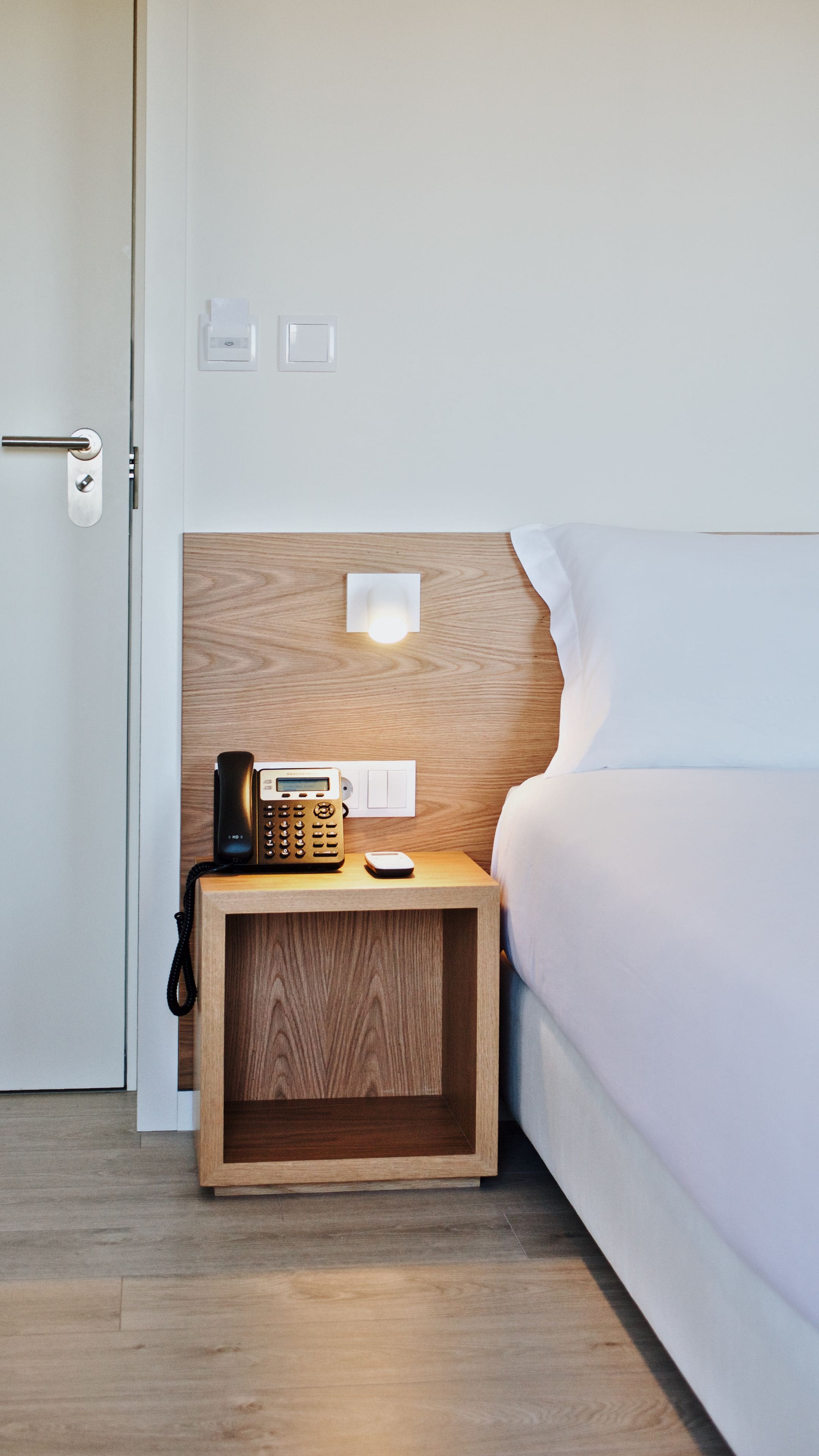 A neatly arranged hotel room with a wooden nightstand, a bedside phone, a wall-mounted light, and a bed with white bedding.