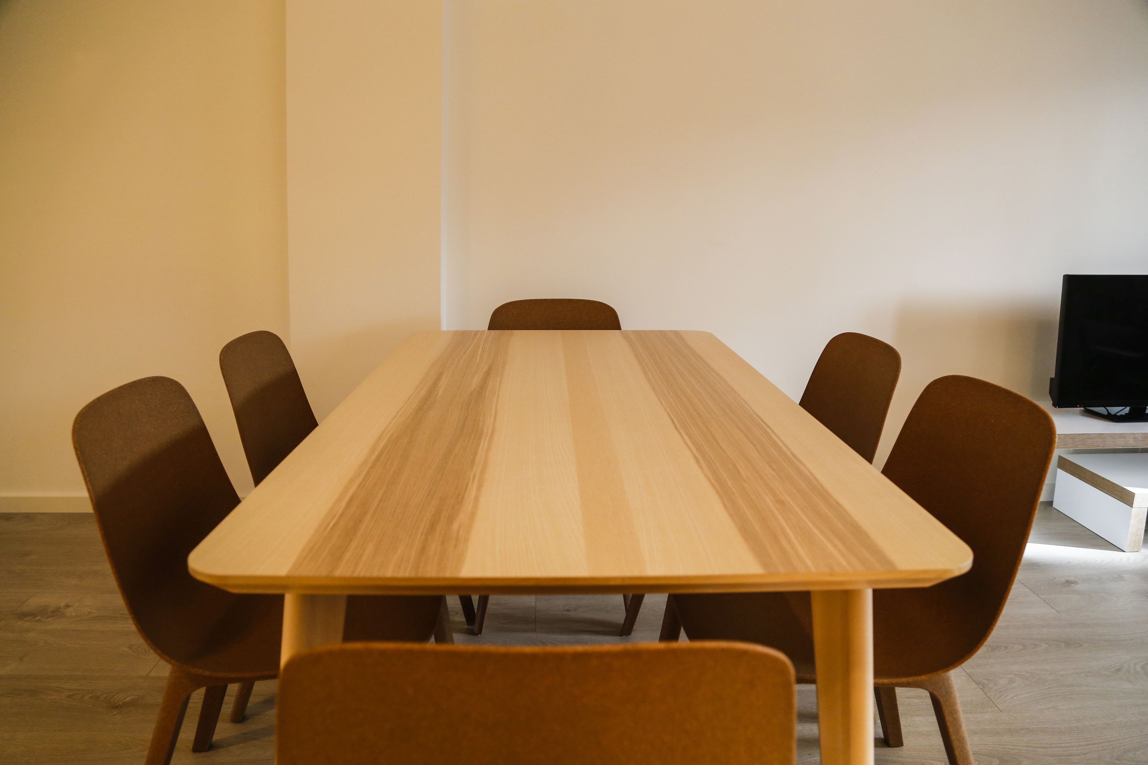 A rectangular wooden table with six chairs, a flat-screen TV on a stand, and a white wall.