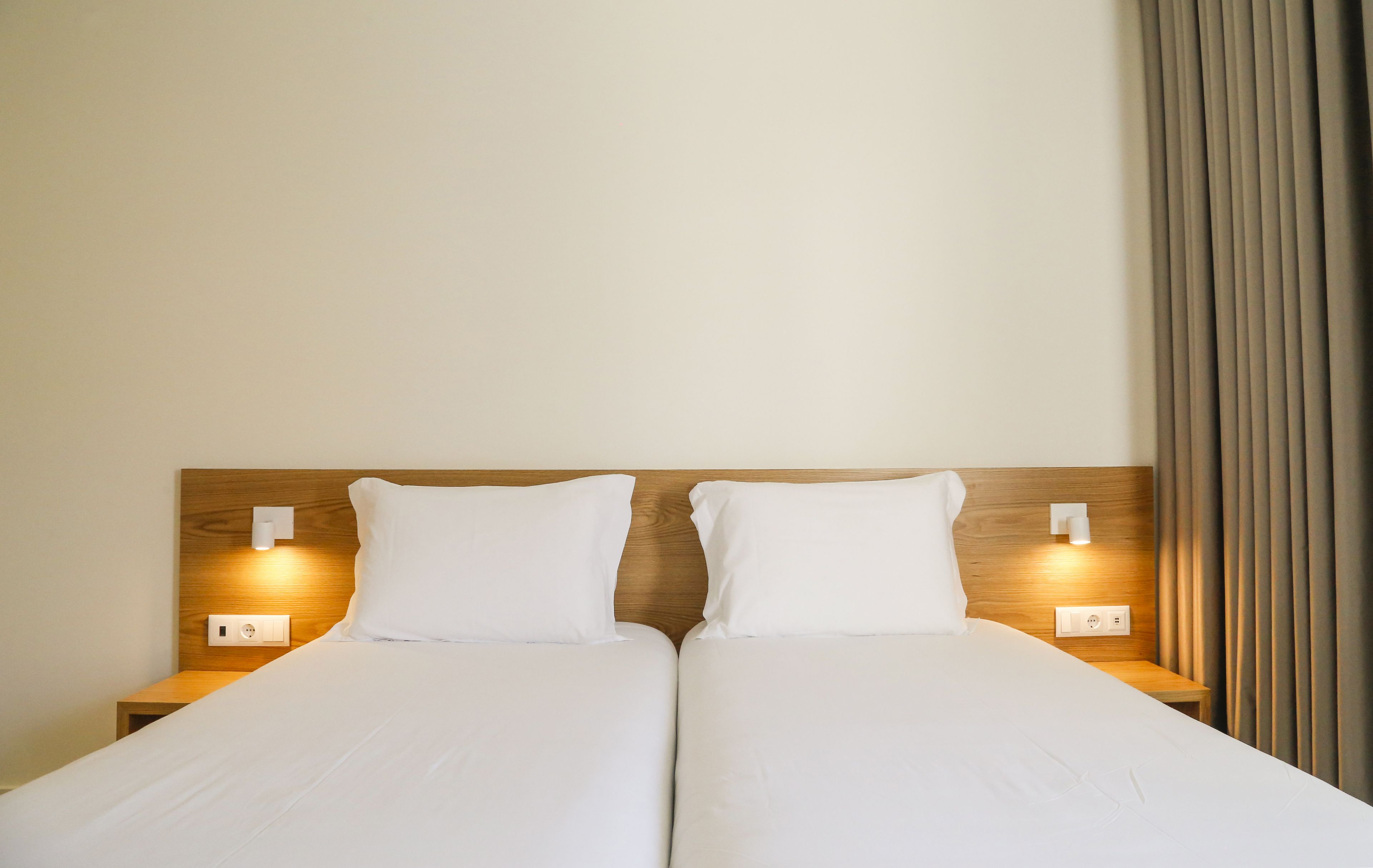 Cama doble con sábanas blancas, cabecera de madera, lámparas de noche y toma de corriente a cada lado.