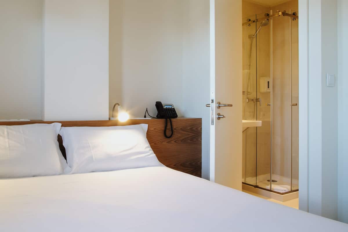 A hotel room with a bed, a bedside phone, and a glass-enclosed shower.
