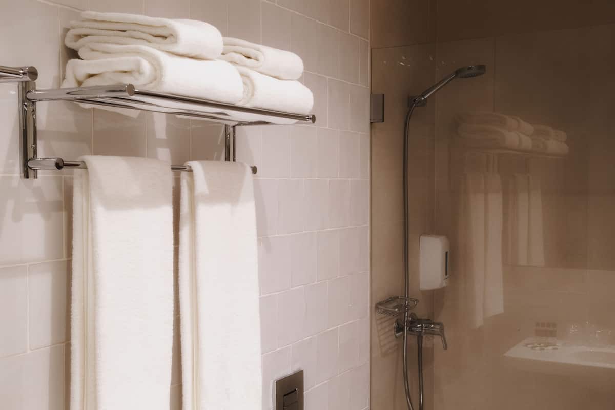 A bathroom with white tiled walls, a glass shower enclosure, a towel rack with folded towels, and a handheld showerhead.