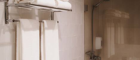 A bathroom with white tiled walls, a glass shower enclosure, a towel rack with folded towels, and a handheld showerhead.