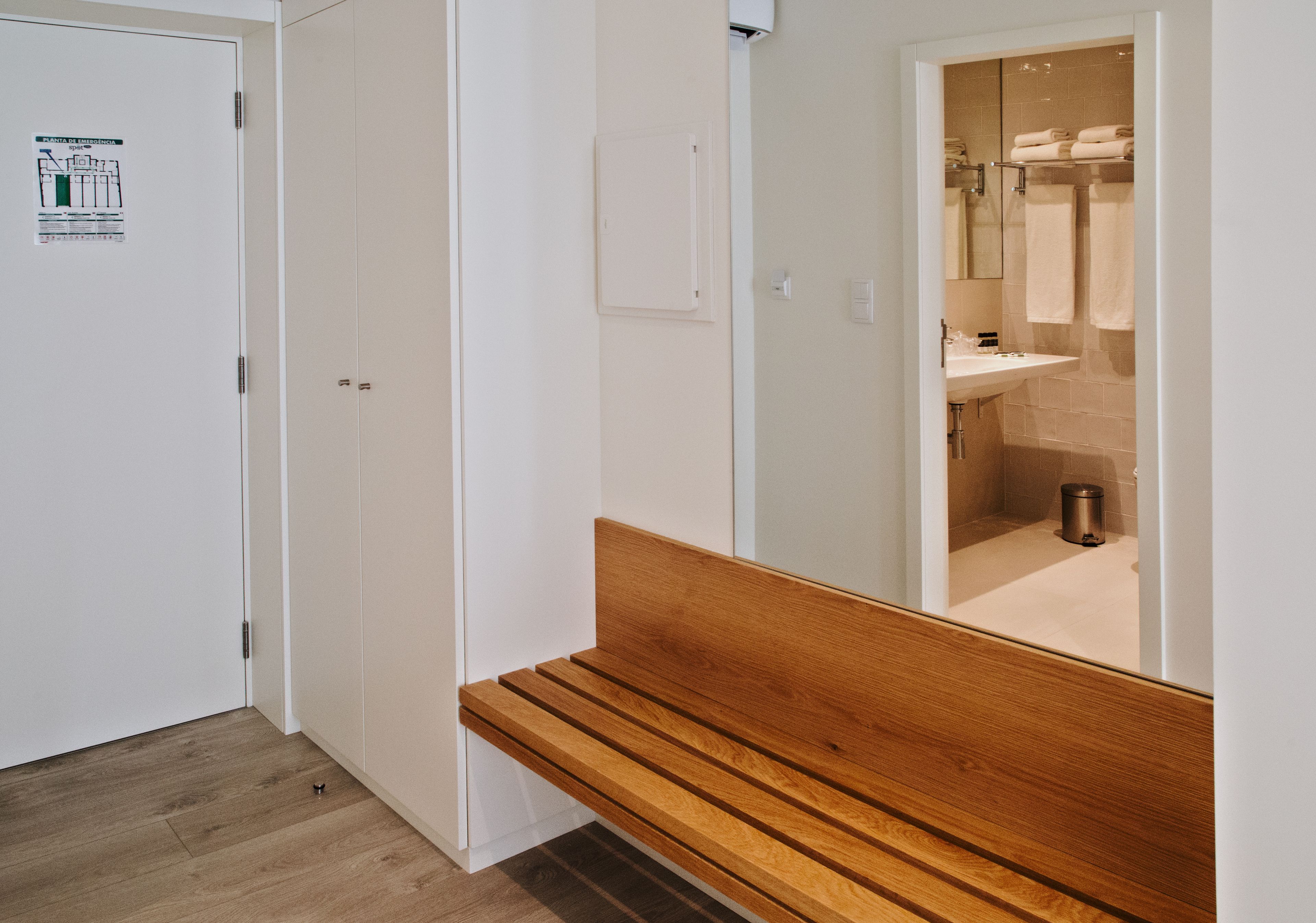 Un pasillo con un banco de madera, una puerta blanca y un espejo que refleja un baño con lavamanos y toallas.