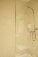 Área de duche com divisória de vidro, chuveiro manual e painel de controlo fixado à parede.