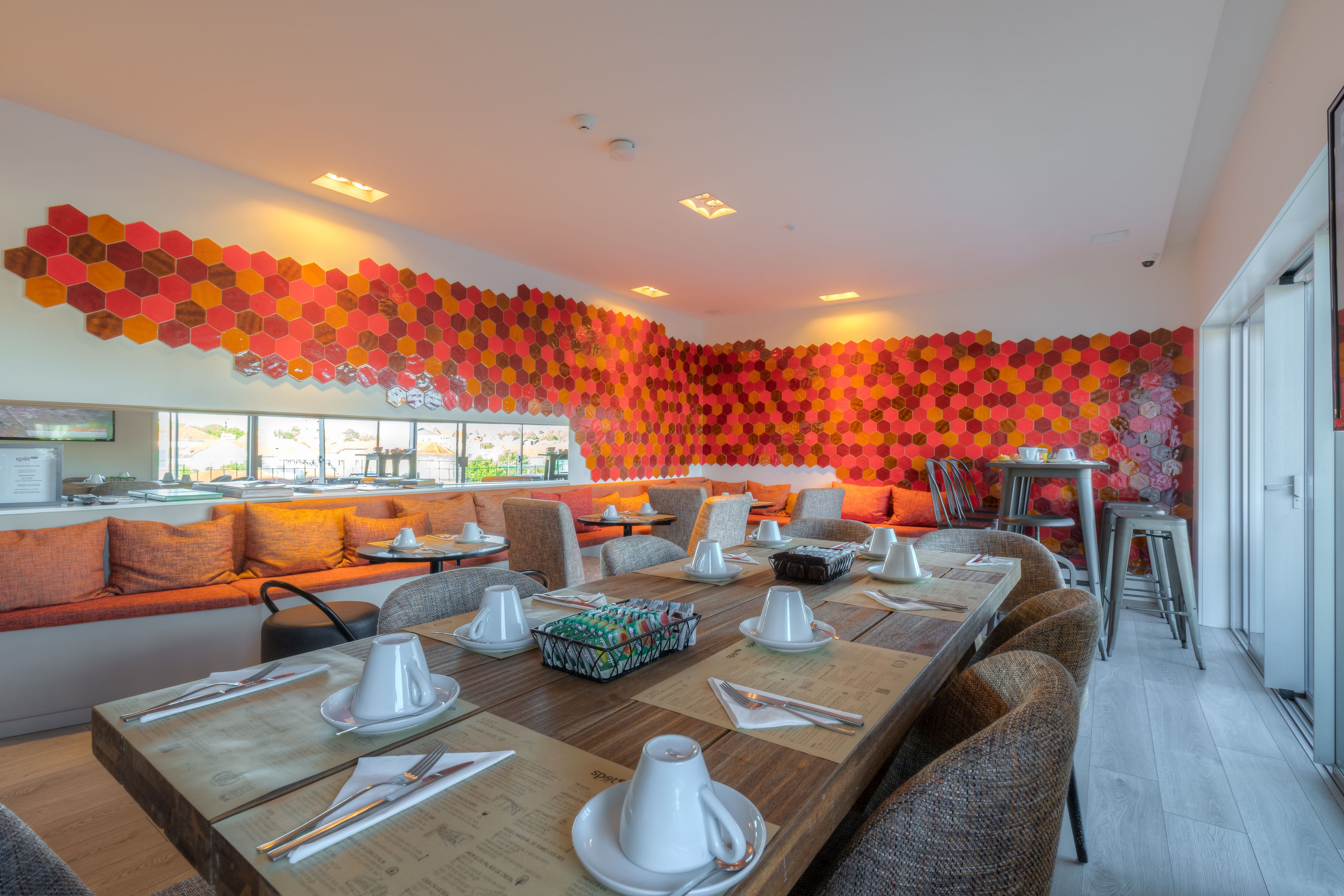 Un restaurant moderne avec une longue table en bois dressée pour le dîner, un mur décoré de motifs hexagonaux aux couleurs vives et un coin salon agrémenté de coussins orange.