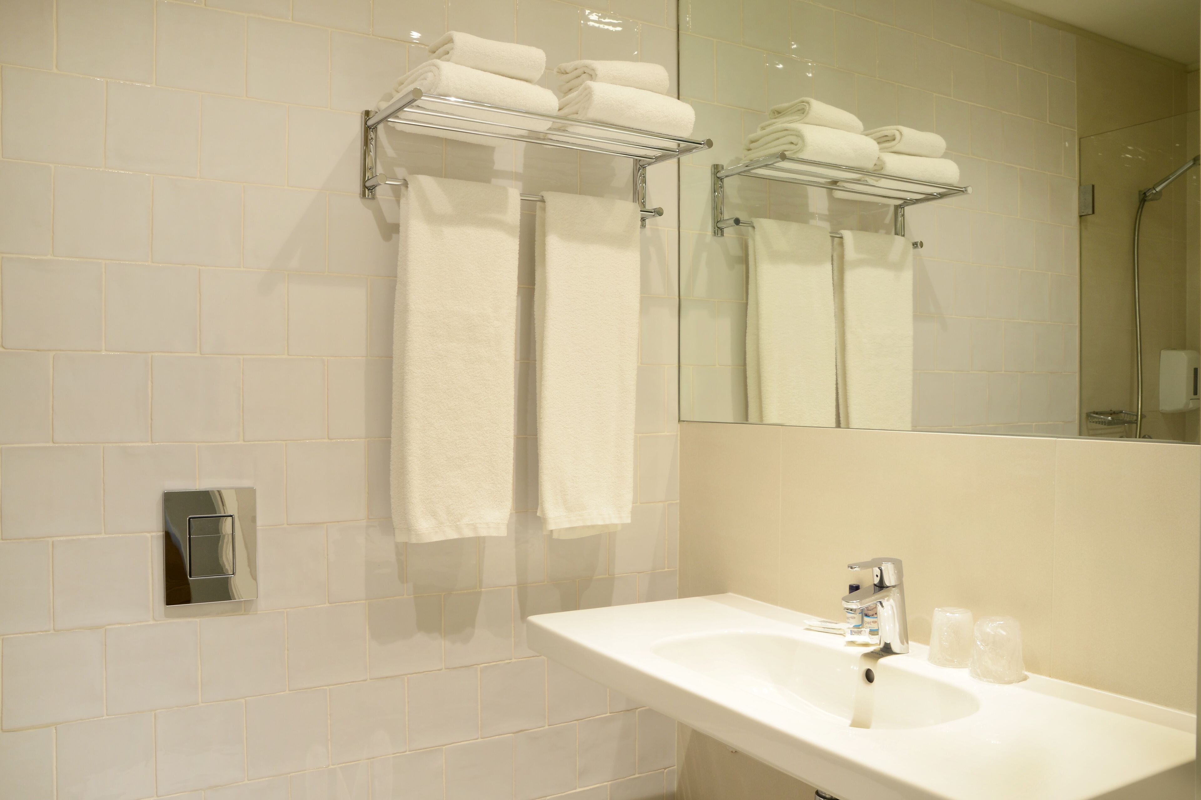 A bathroom with white tiled walls, a sink with a mirror, and two towel racks holding folded towels.