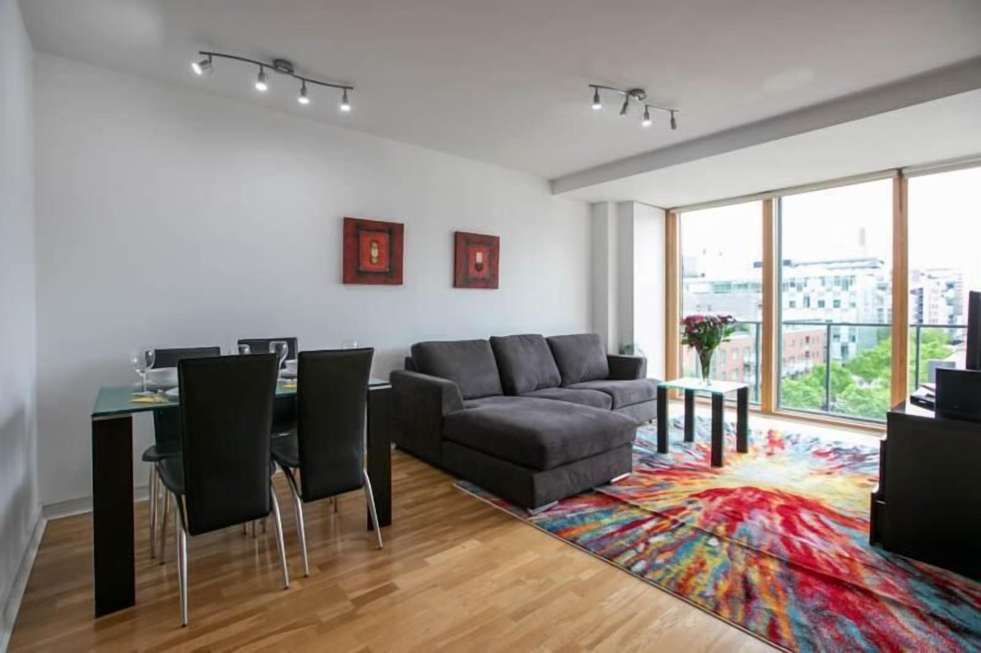 A modern living room with a dining area, large windows, and a colorful rug.