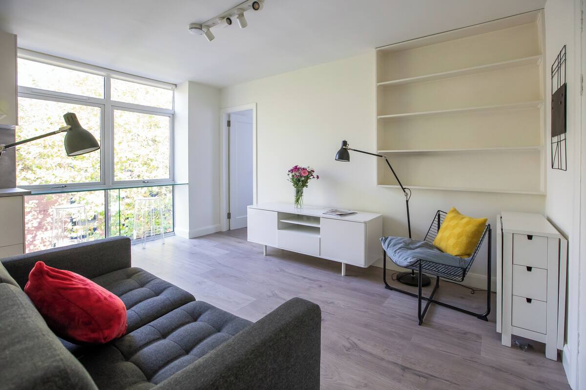Ein modernes Wohnzimmer mit grauem Sofa, rotem Kissen, weißem Konsolentisch mit Blumenvase, einer schwarzen Stehlampe und einem weißen Schrank.