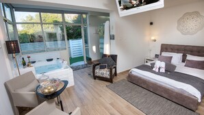 Ein modernes Hotelzimmer mit einem großen Bett, einer Sitzecke mit Tisch, einer kleinen Couch und Blick auf einen Garten durch eine Schiebetür.
