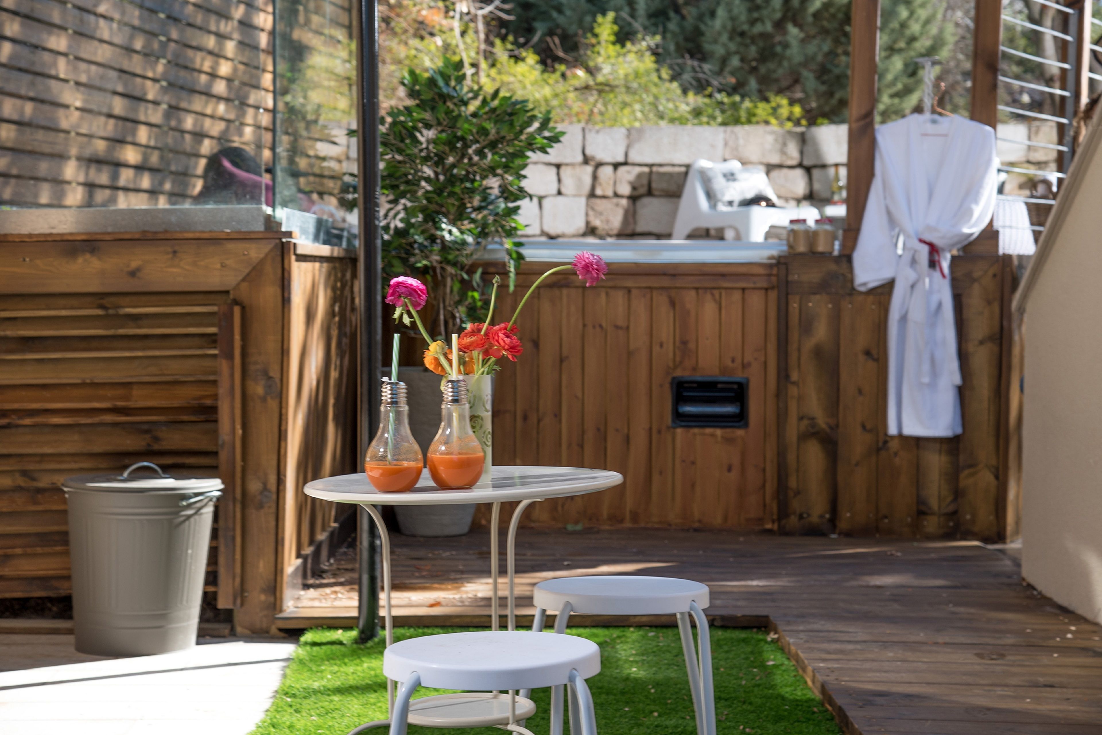 A wooden deck with a small table and two stools, a trash can, and a white robe hanging on a rack.