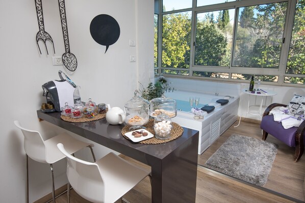 A modern kitchen with a dark countertop, white chairs, and a view of trees through large windows. - Osnata Boutique Suites (Kfar Vradim)