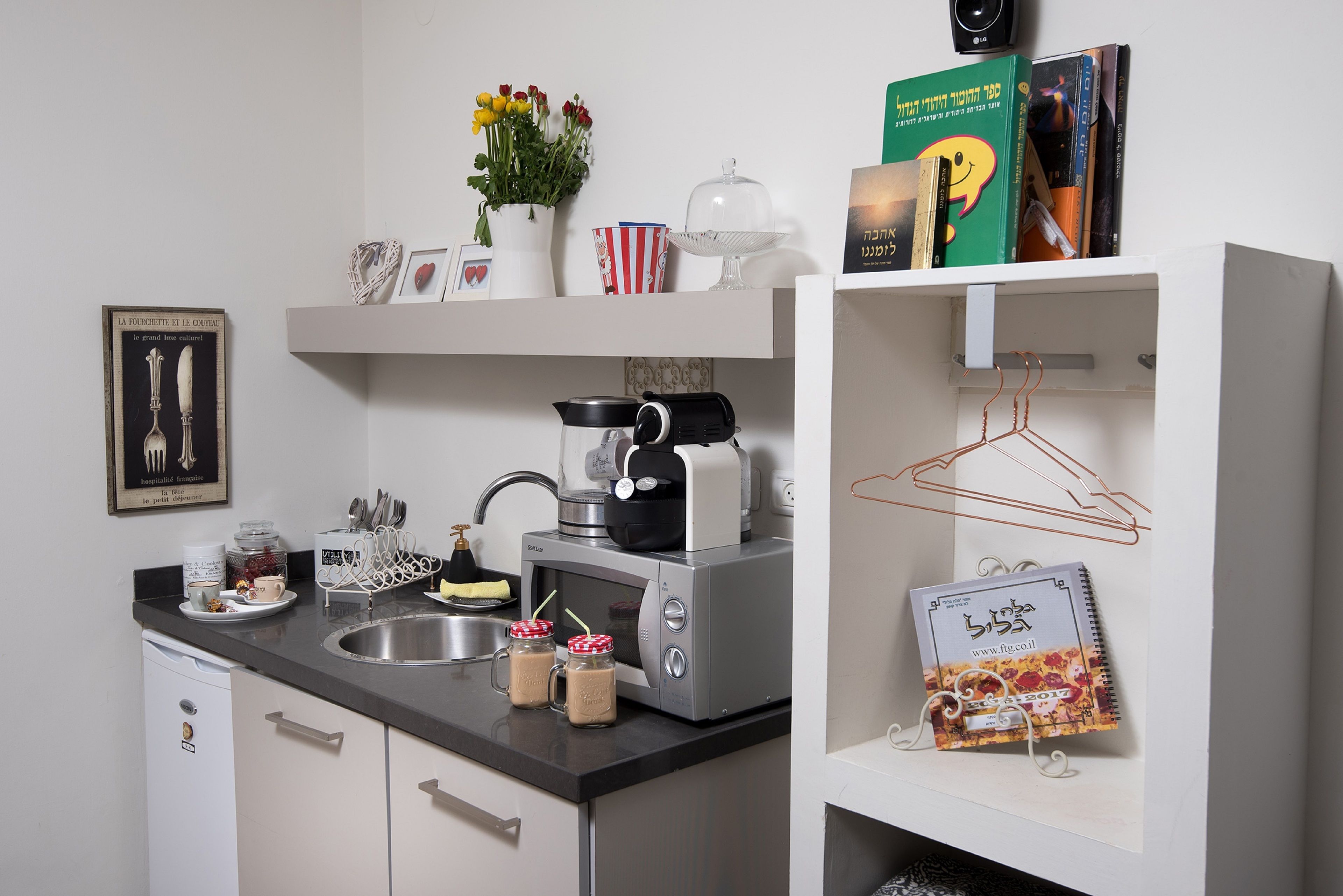 Una cocina compacta con microondas, tostadora y lavamanos. Hay elementos decorativos como un florero con flores, un cuadro enmarcado y un calendario.