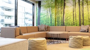 A modern living room with a large window, a curved sofa, a small round table, and a forest mural on the wall. - Oslo Hostel Rønningen (Oslo)