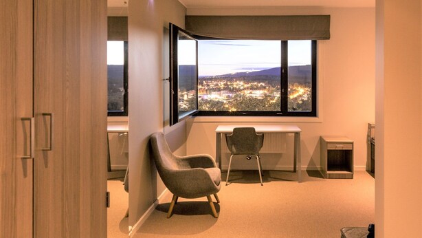 A modern room with a large window offering a city view, a desk, a chair, and a small pet bed. - Oslo Hostel Rønningen (Oslo)