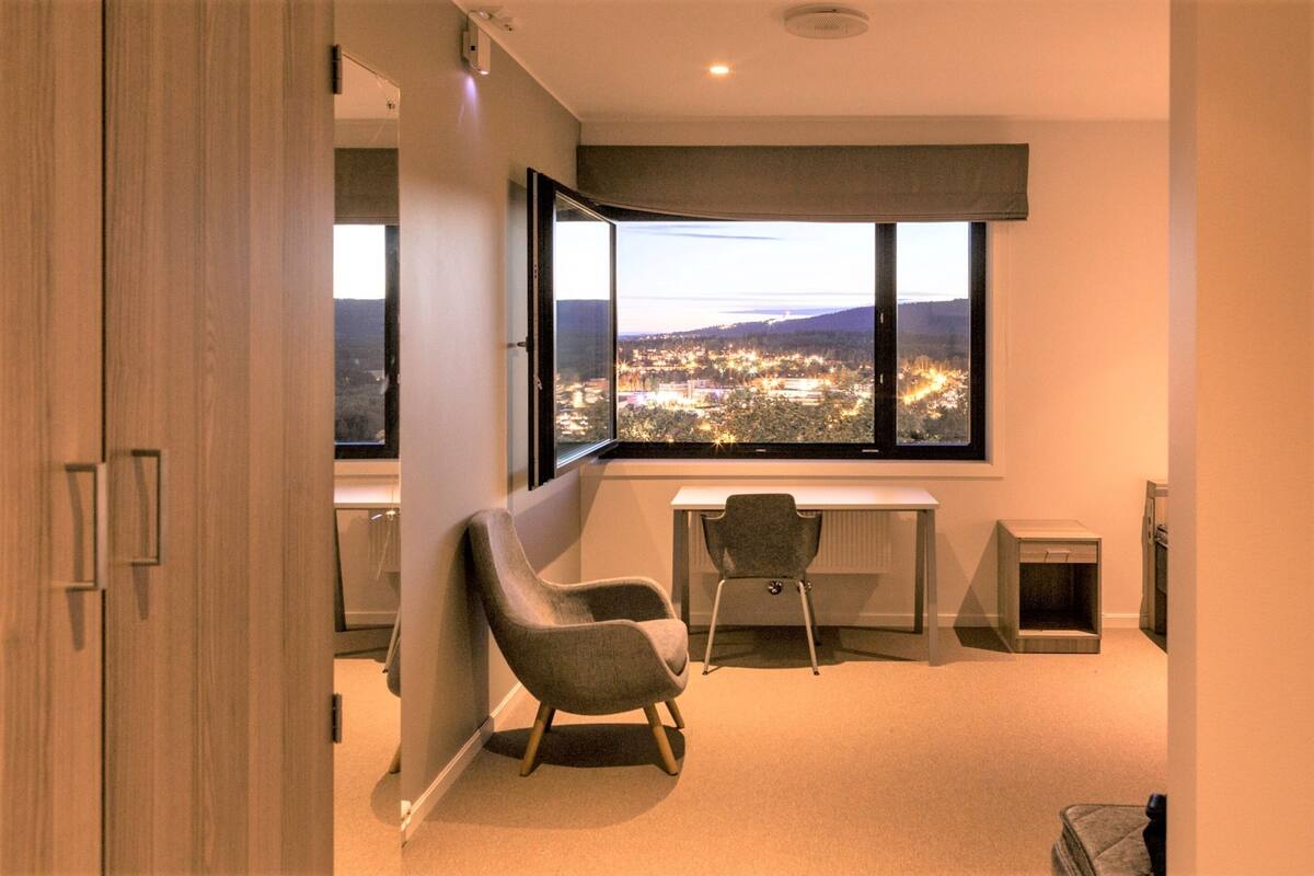 A modern room with a large window offering a city view, a desk, a chair, and a small pet bed.