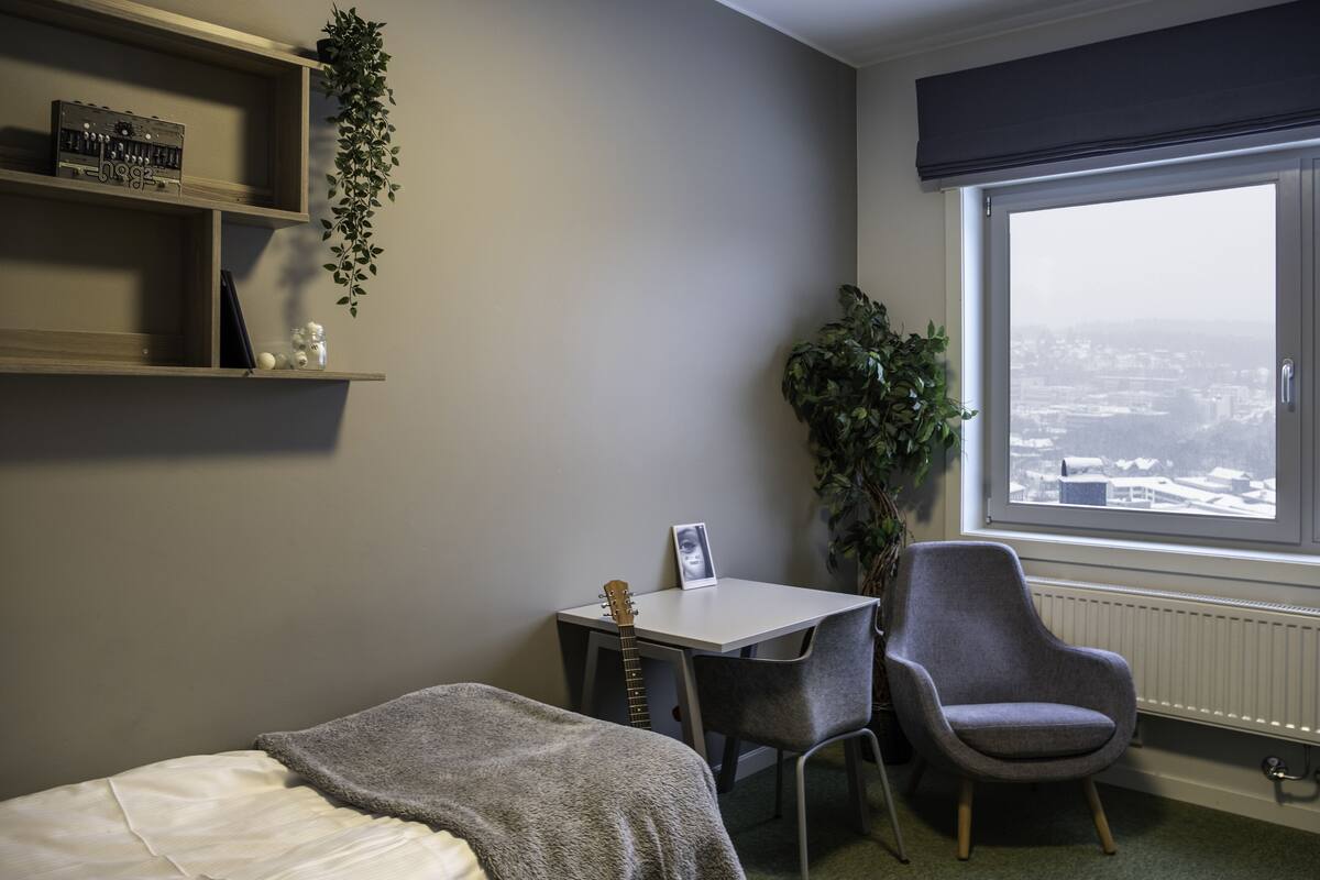 A bedroom with a bed, a desk, a chair, a radiator, and a window with a view of a cityscape.