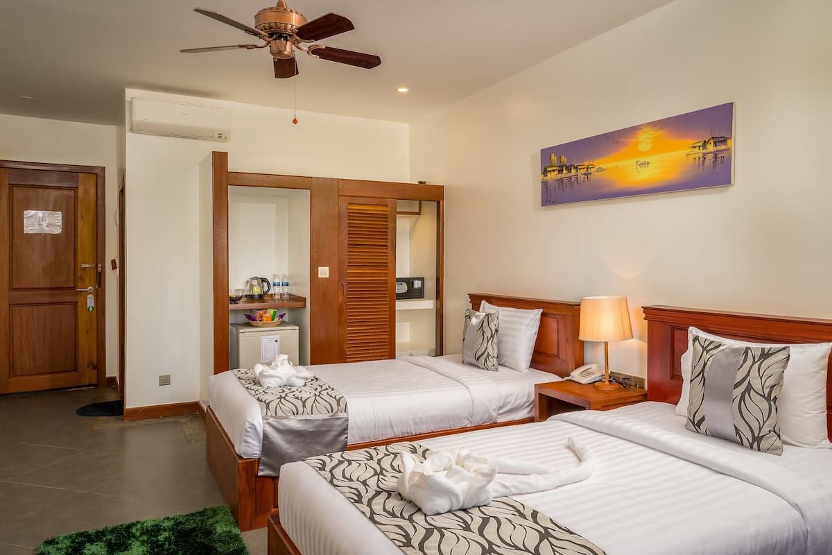 A hotel room with two beds, a ceiling fan, a painting of a sunset, and a small table with a lamp.