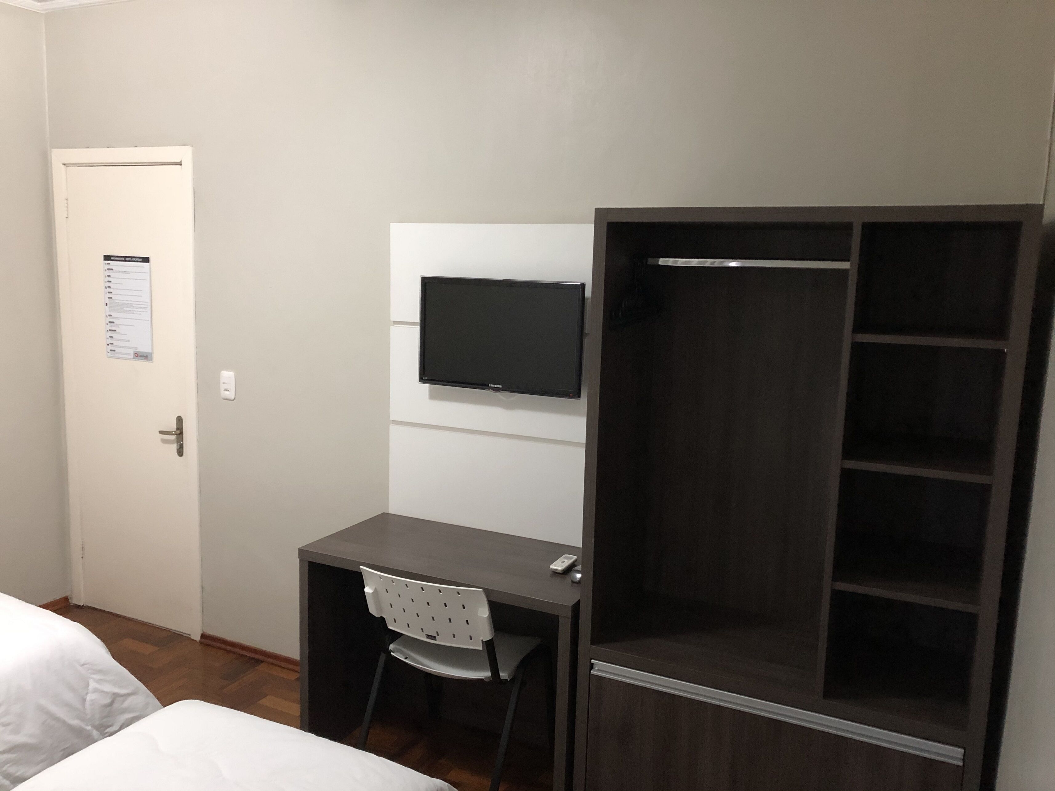 Quarto de hotel com cama, escrivaninha, cadeira, televisão e guarda-roupa.