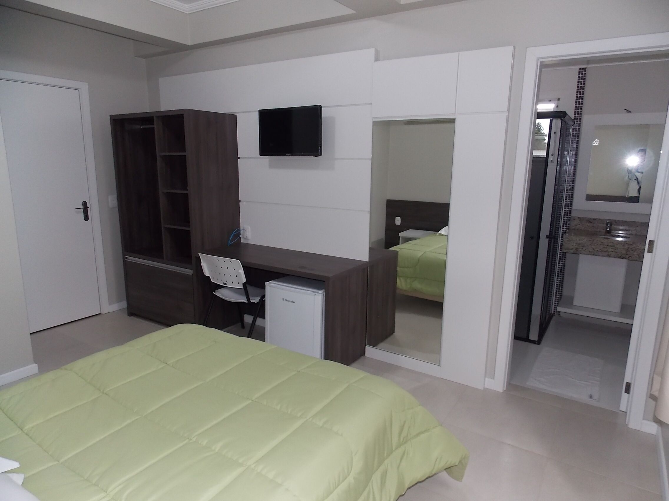 A bedroom with a bed, a desk, a chair, a wardrobe, a television, and a bathroom visible through an open door.