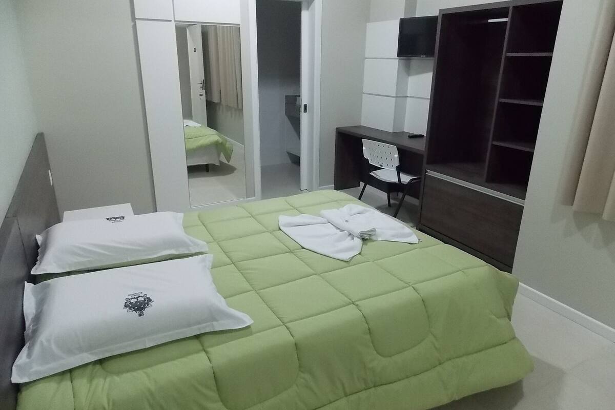 A neatly made bed with a green bedspread and white pillows, a desk with a chair, a wardrobe, and a mirror.