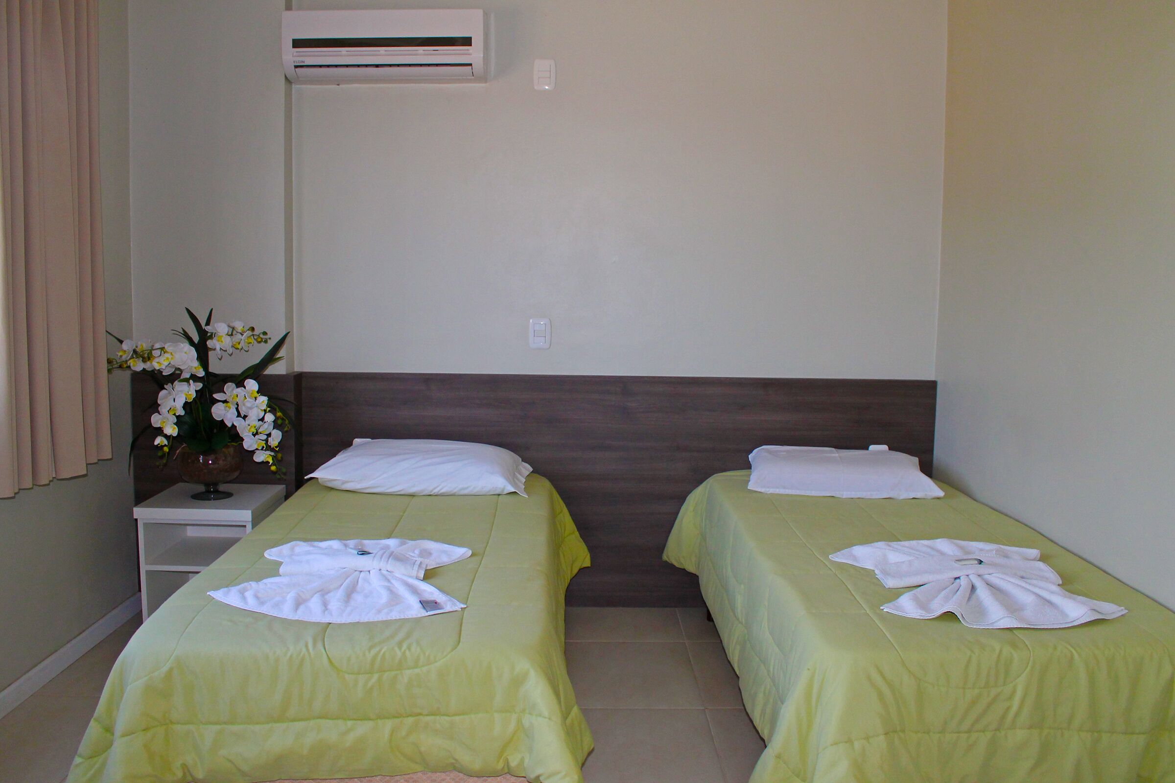 Habitación de hotel con dos camas, cada una con cubrecama verde y almohadas blancas. Hay una mesita con un jarrón de flores y una toalla blanca.