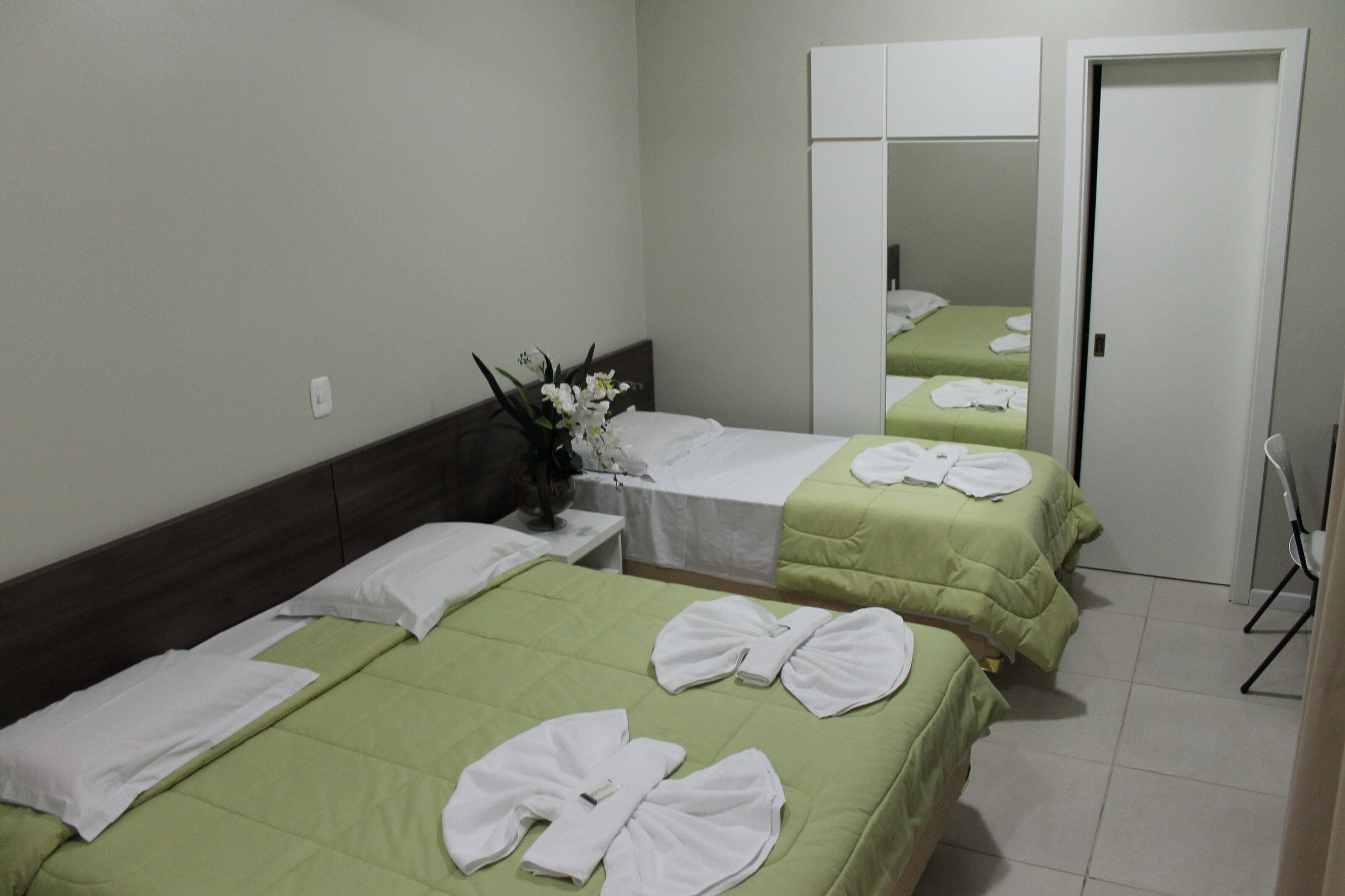 A hotel room with two beds, each with green bedding and white towels. There is a small table with a vase of flowers and a mirror on the wall.