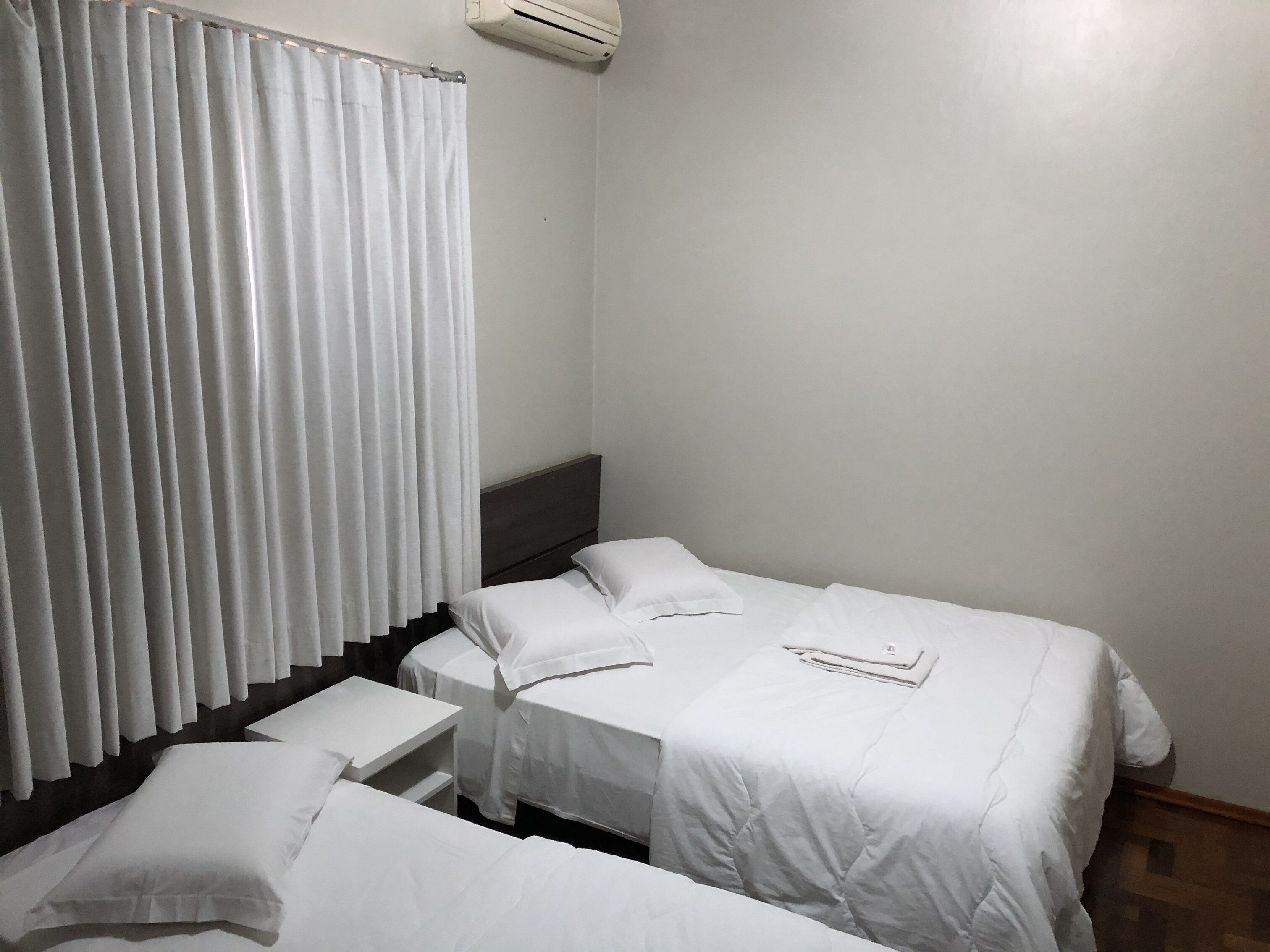 A small, clean room with two beds, a small table, and a window with white curtains.