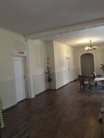 Una habitación amplia con suelo de madera, paredes blancas y un candelabro. Hay una mesa de comedor con sillas y una planta en maceta.