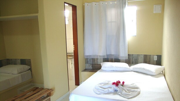 Quarto individual com uma cama, um banco de madeira, um banheiro visível por uma porta e uma janela com cortina.