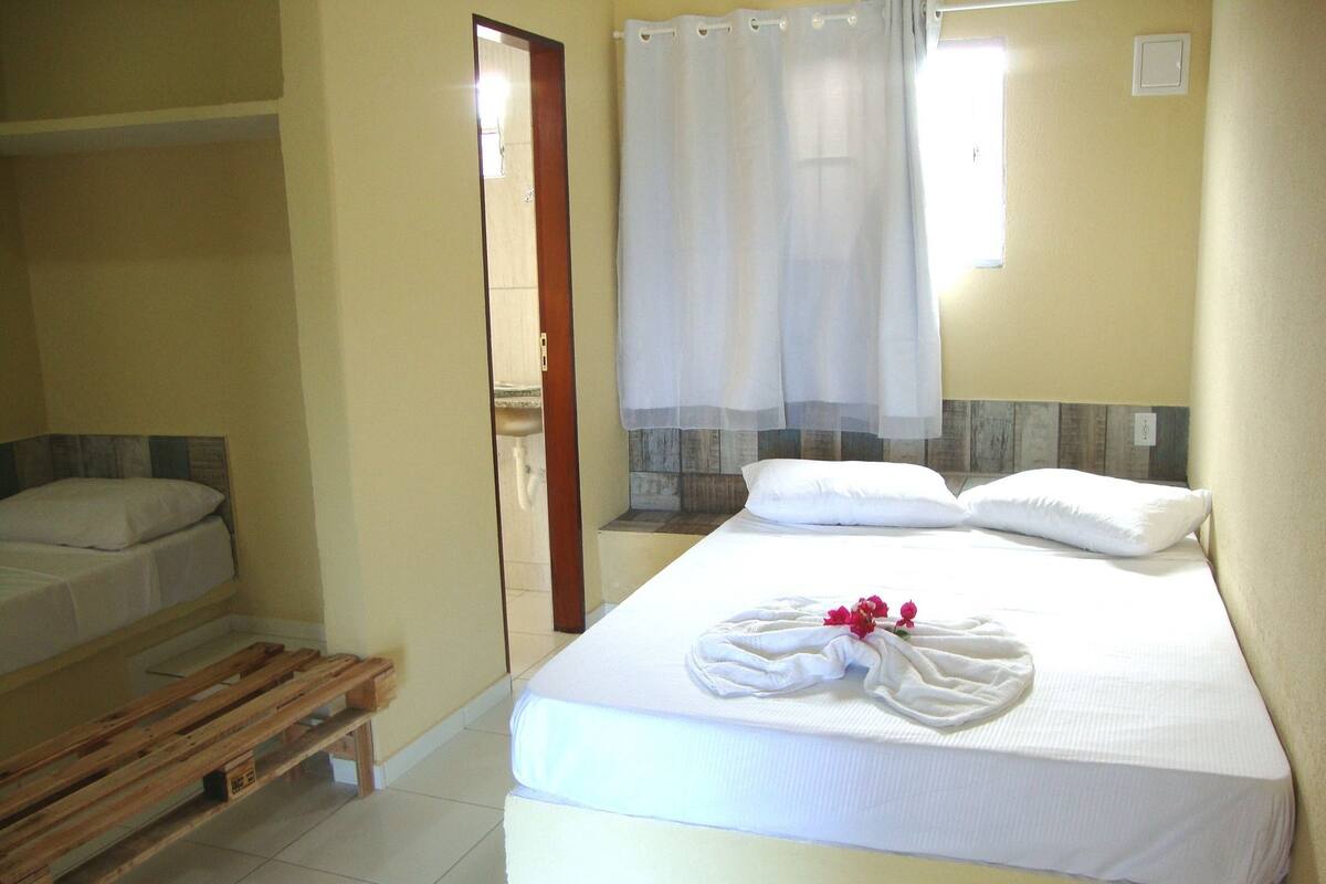 Ein Zimmer mit einem Einzelbett, einer Holzbank, einem durch eine Tür einsehbaren Badezimmer und einem Vorhang vor dem Fenster.