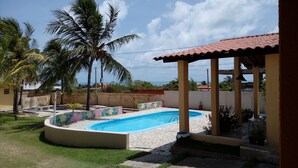Piscina com deck ladrilhado, rodeada por palmeiras e um espaço coberto com varanda.