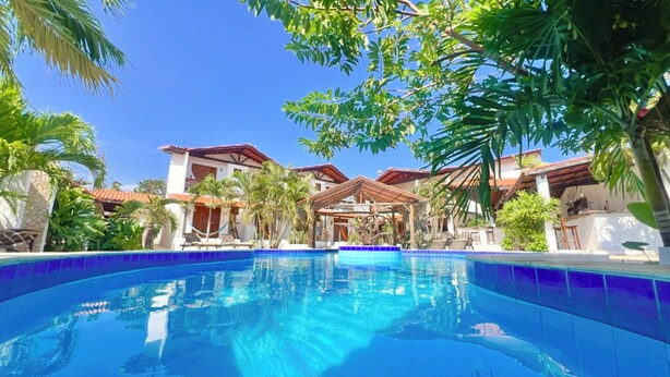 A swimming pool surrounded by palm trees and a building with a red roof. - Hotel Pousada 360° (Paracuru)