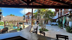 An outdoor patio with a pool, wooden tables, and chairs, surrounded by buildings with red-tiled roofs and a thatched roof structure. - Hotel Pousada 360° (Paracuru)