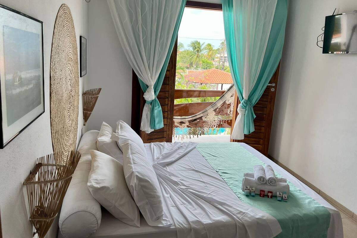 A hotel room with a bed, white curtains, a hammock, and a view of a pool and palm trees.