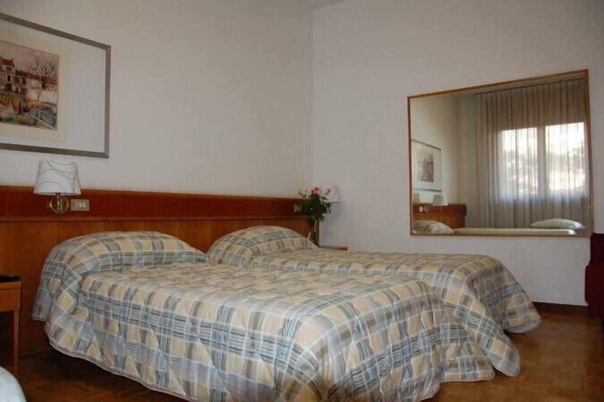 A room with two beds, a wooden headboard, a bedside table with a lamp, a mirror, and a framed picture on the wall.
