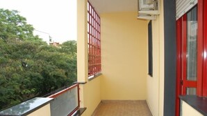 Een smal balkon met een betegelde vloer, een rode deur en een raam met een metalen traliewerk.