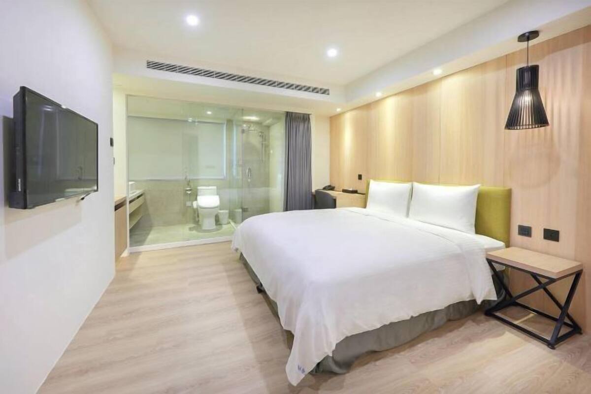 Une chambre d’hôtel moderne équipée d’un lit, d’une table de chevet, d’une télévision et d’une douche séparée par des parois en verre.