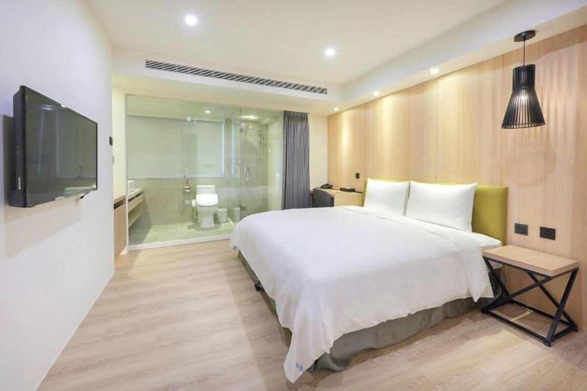 Ein modernes Hotelzimmer mit Bett, Nachttisch, Fernseher und einer gläsernen Duschkabine.