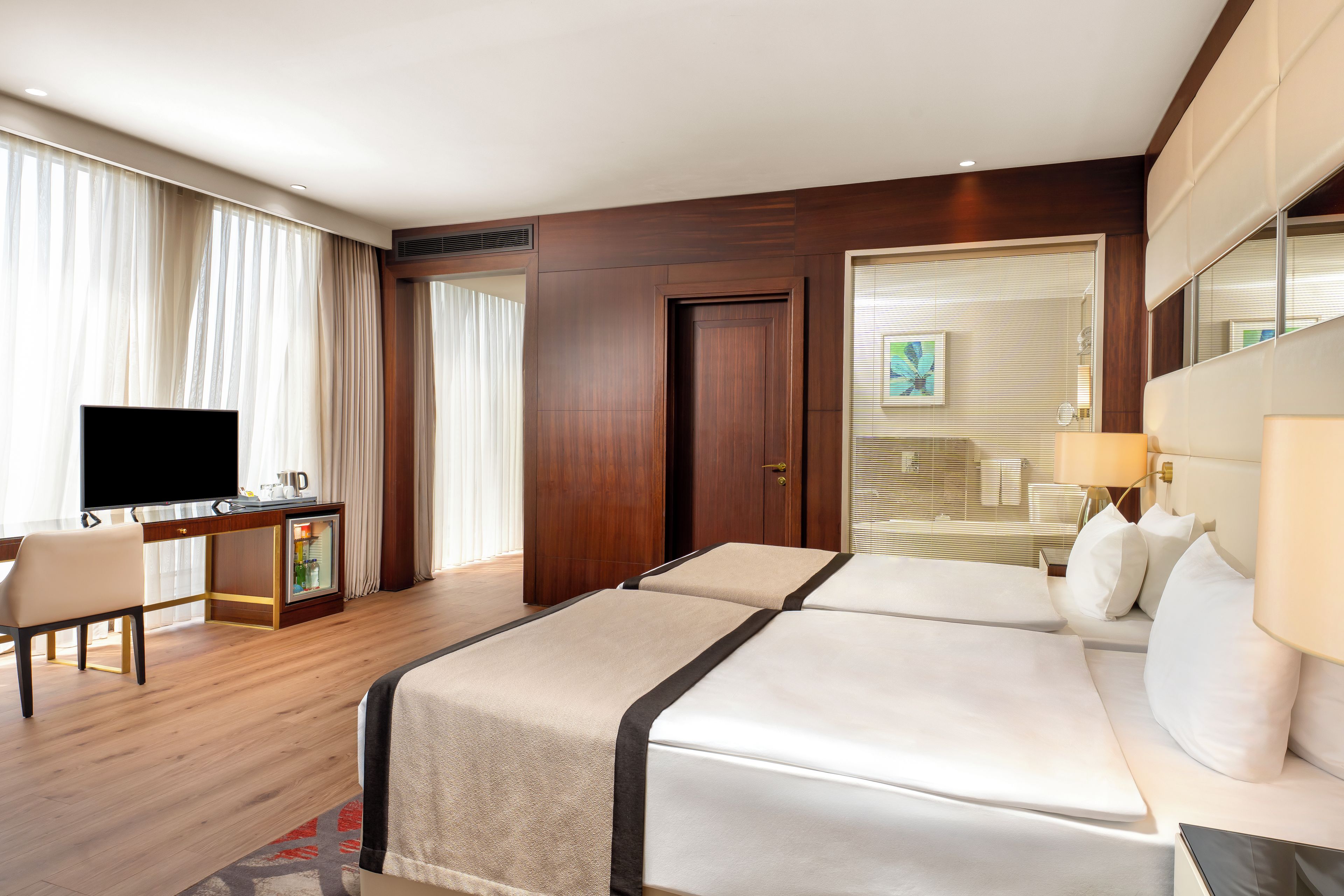A hotel room with a bed, a desk with a television, a minibar, and a large window with curtains.