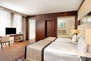 A hotel room with a bed, a desk with a television, a minibar, and a large window with curtains.