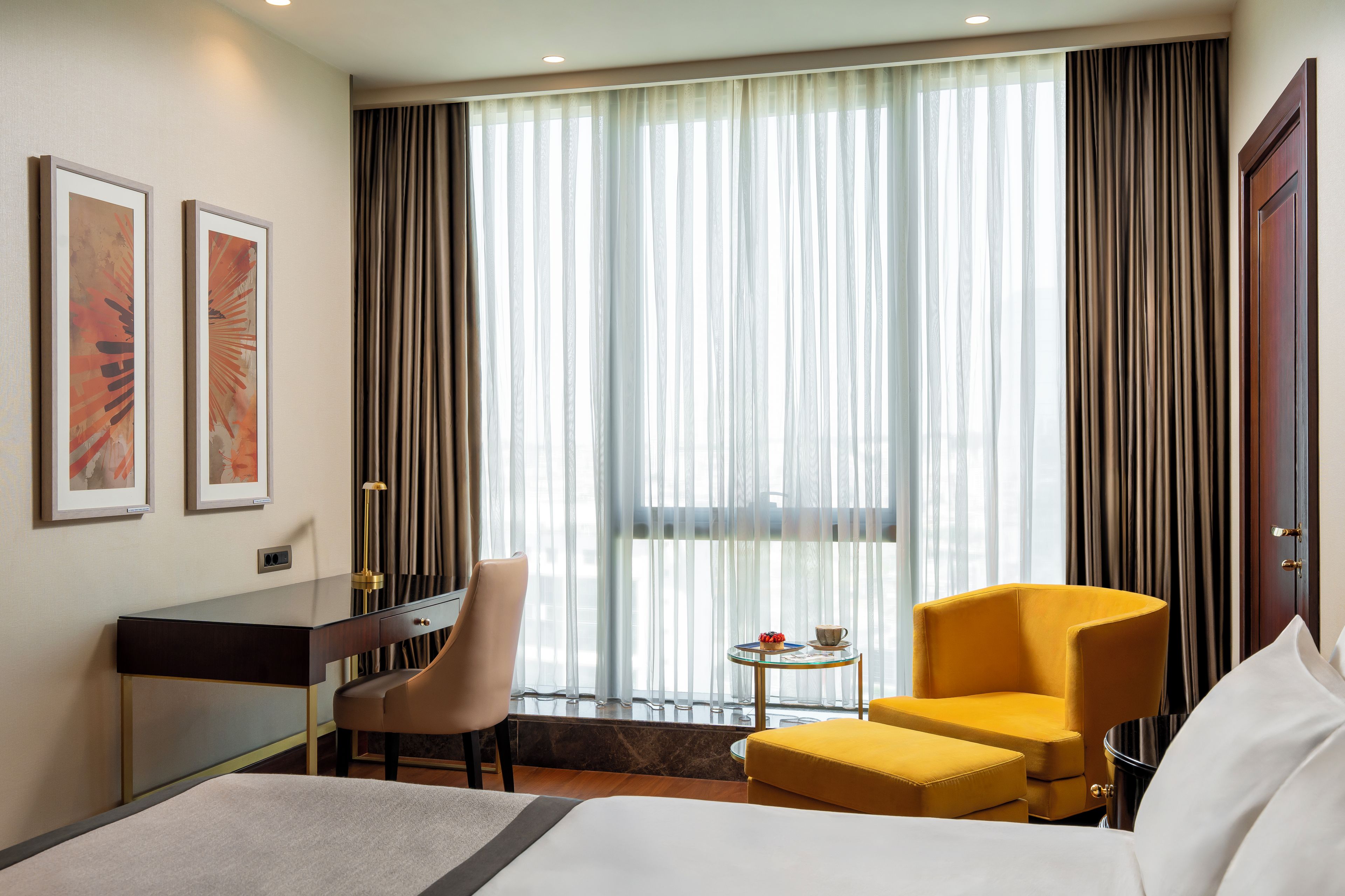 A hotel room with a bed, a desk, a chair, a small table with a cup and a plate, and a window with curtains.
