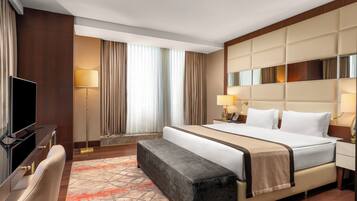 Habitación de hotel con cama, mesitas de noche, escritorio con televisor de pantalla plana, banca y un amplio ventanal con cortinas.