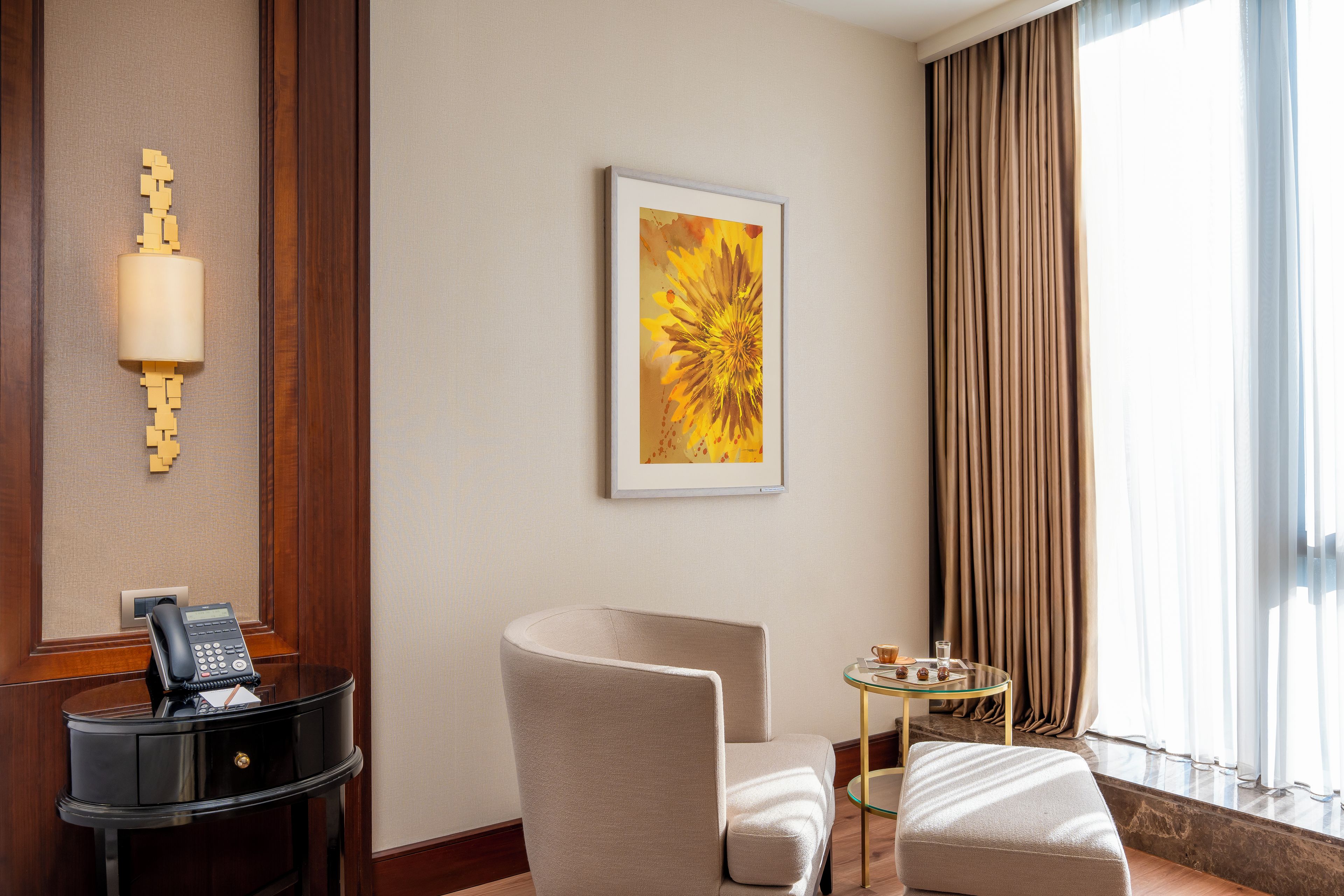 Una habitación de hotel con escritorio, teléfono, silla, reposapiés y un cuadro en la pared.