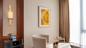 A hotel room with a desk, telephone, chair, ottoman, and a painting on the wall.