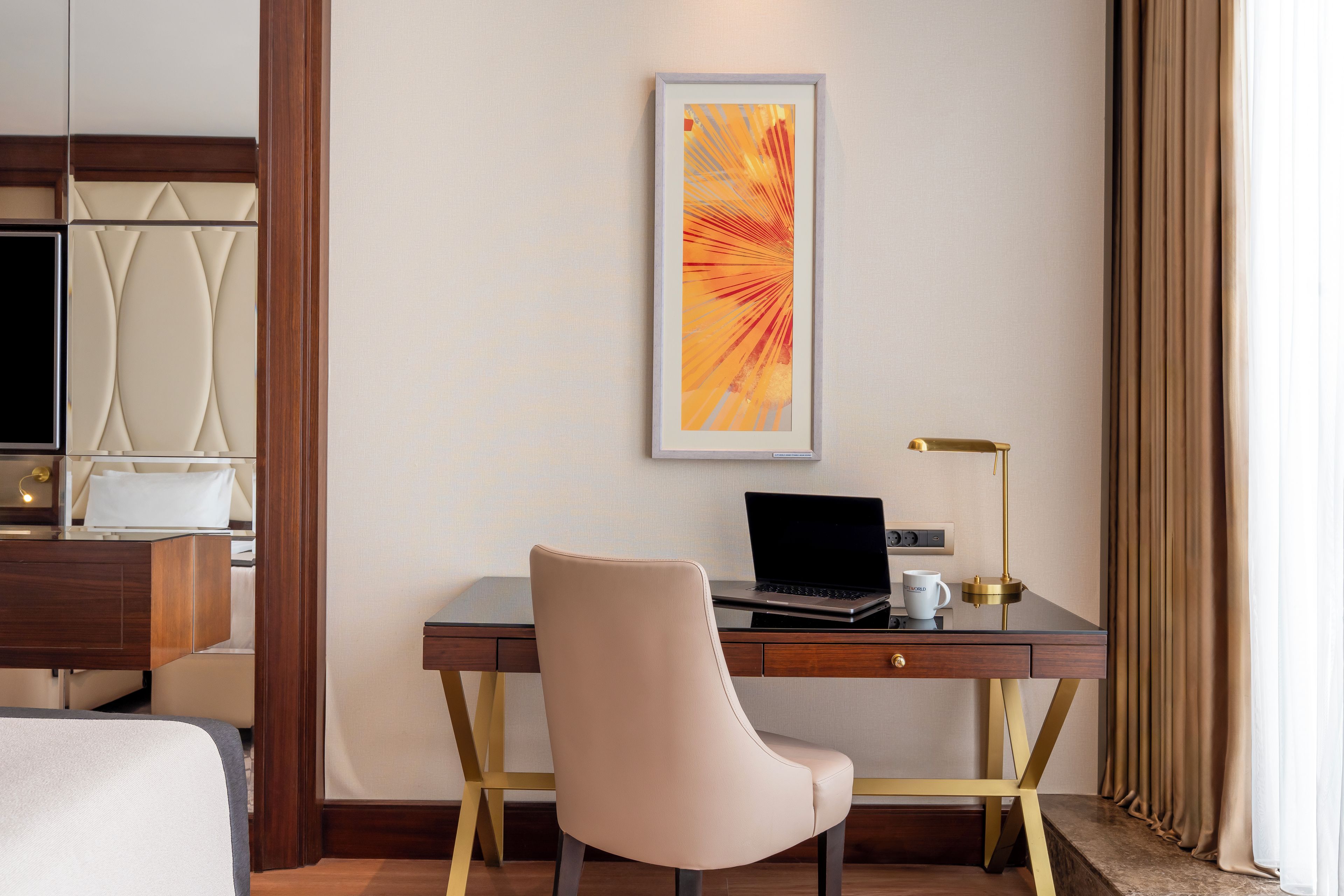 A hotel room with a desk, a laptop, a chair, a bedside table, a lamp, and a bed.