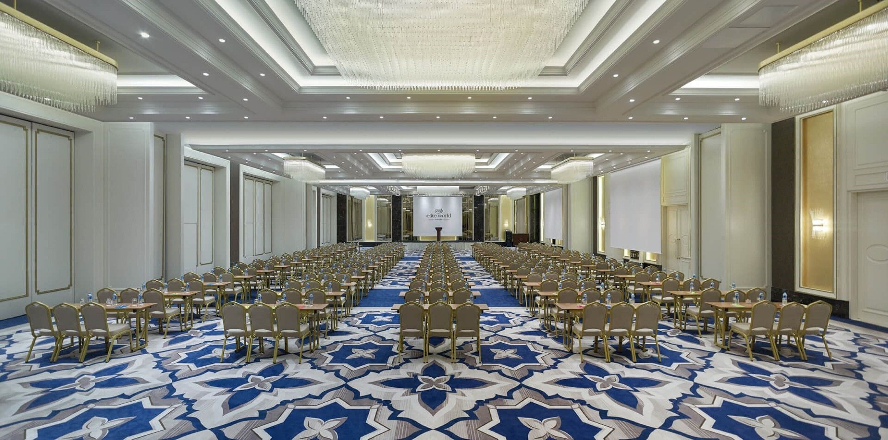 A large conference room with rows of tables and chairs, a patterned carpet, and a chandelier.