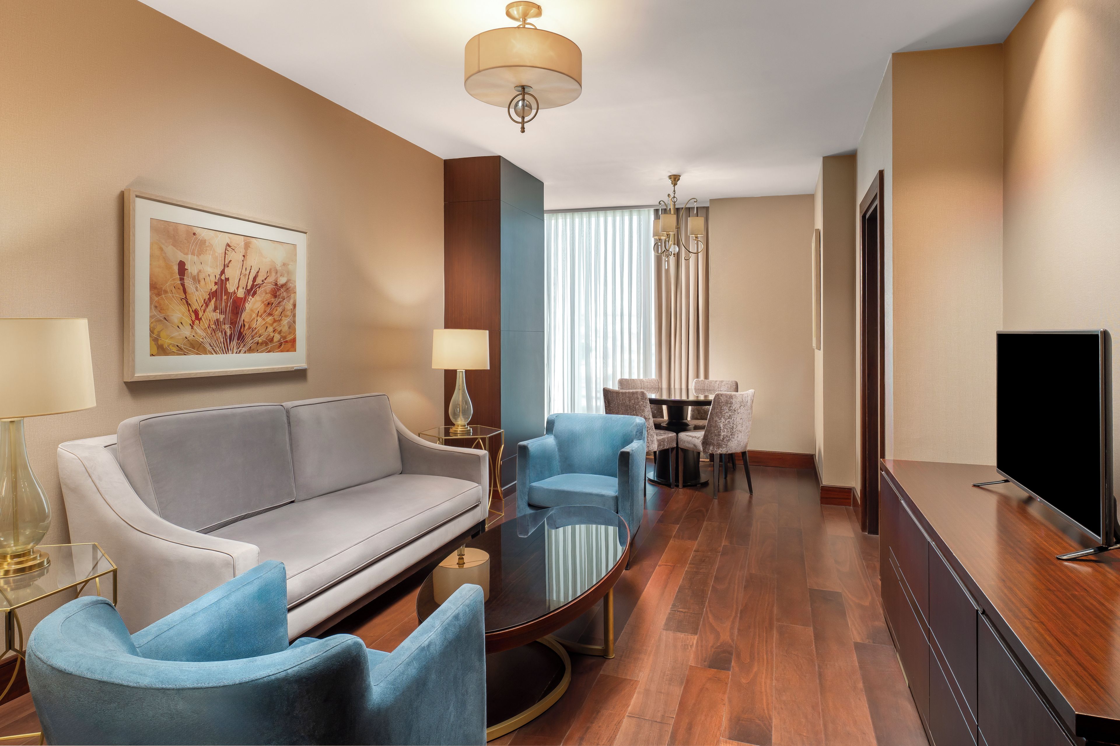 A hotel room with a sofa, armchairs, a TV, a dining table with chairs, and a chandelier.