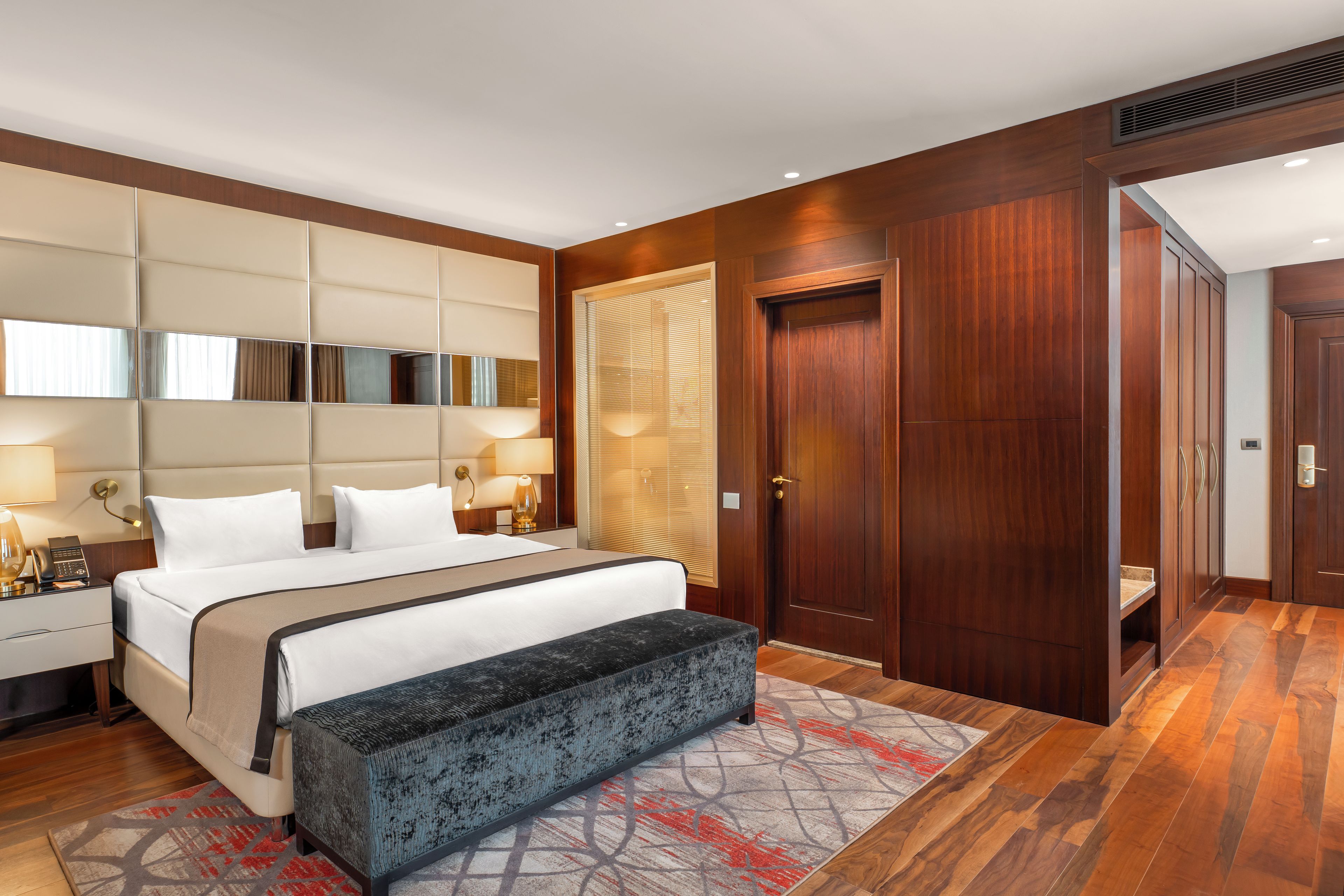 Een hotelkamer met een groot bed, bedlampjes, een bankje, een spiegel, een deur en een badkamer zichtbaar door een raam.