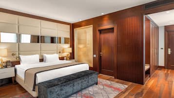Een hotelkamer met een groot bed, bedlampjes, een bankje, een spiegel, een deur en een badkamer zichtbaar door een raam.